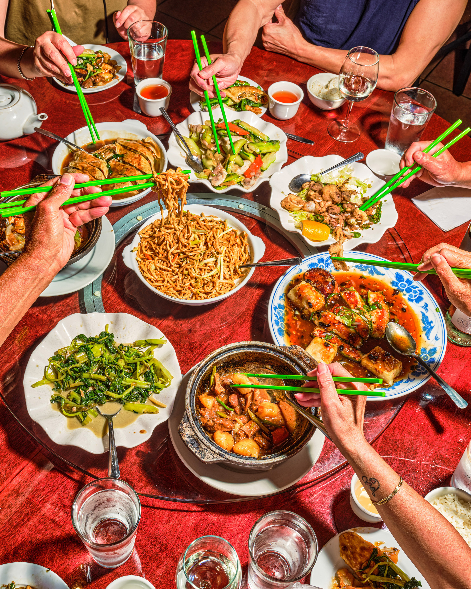 Overhead view of a family style table at Uncle Lou: noodles, stir fries and salt and pepper seafood on a red lazy susan, hands reaching in with green chopsticks